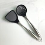 A modern Honglida silicone wok skimmer with a black head and stainless steel handle, resting in a nonstick pan.
